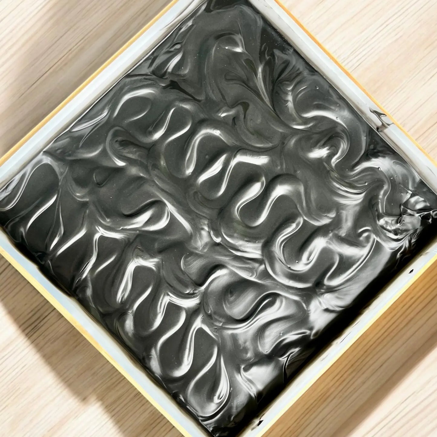 Un contenant carré est rempli d'un d'un savon exfoliant. Les tourbillons créent un motif ondulé à la surface. Rappelant le Savon exfoliant ALI au charbon actif de La Sultana Savonnerie | Savons et soins naturels faits main | Québec, cela pourrait être parfait pour les peaux à tendance grasse. Le contenant repose sur une surface en bois clair.