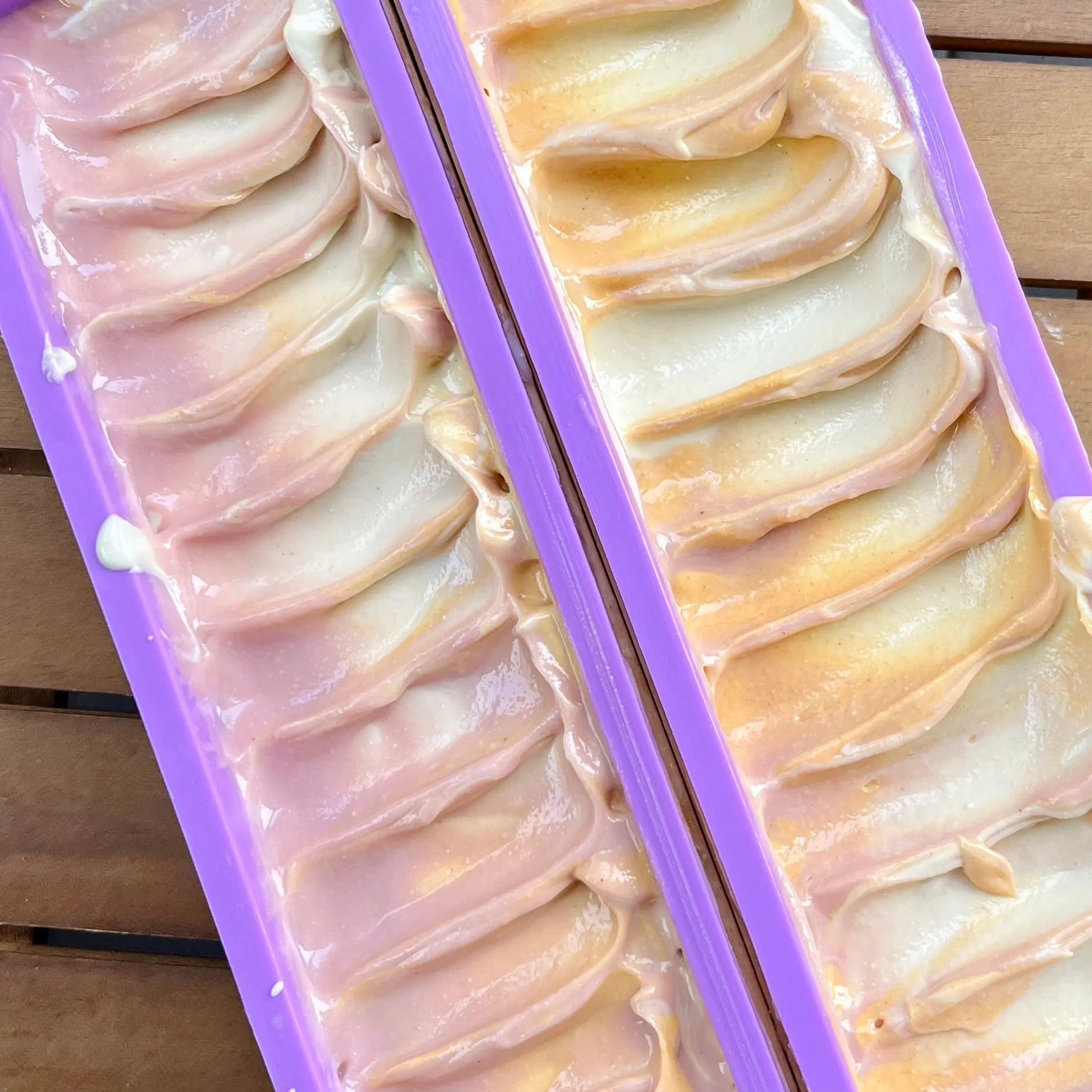 Vue de dessus de deux plateaux de mélange de Savon au lait de coco SULTANE argile blanche et pêche dans des moules, avec des couleurs pastel tourbillonnantes allant du violet clair au pêche. Les moules sont violets et quelques bulles et déversements sont visibles sur la surface en bois en dessous. Ce savon artisanal est fabriqué par La Sultana Savonnerie | Savons et soins naturels faits main | Québec.