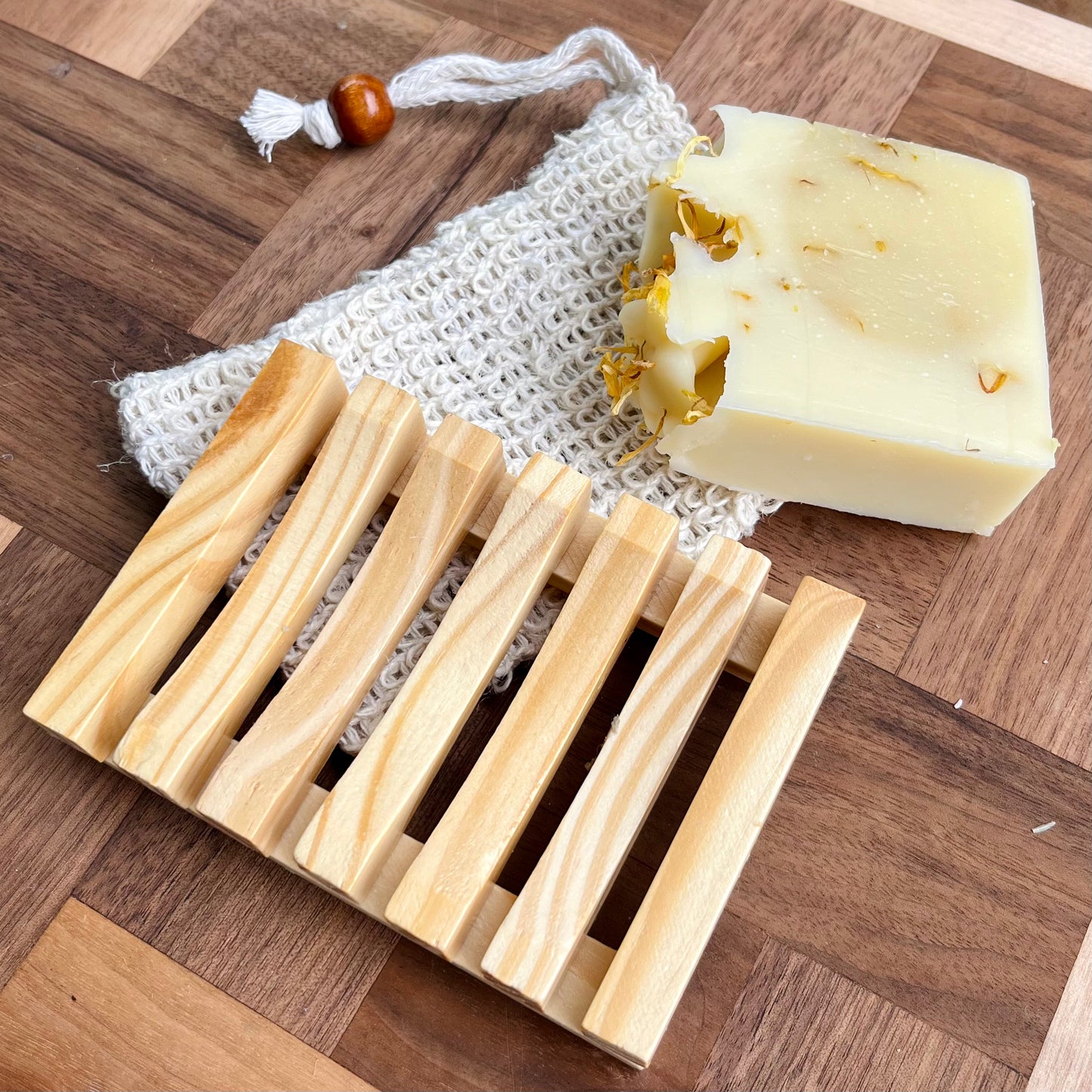 Un Porte-savon et pochette en sisal de La Sultana Savonnerie, contenant une barre de Savons artisanaux, est astucieusement placé sur une surface en bois. Le savon présente une texture crémeuse avec des pétales de fleurs séchées incrustés. Le sac à gommage en coton est agrémenté d'une perle en bois sur son cordon de serrage.