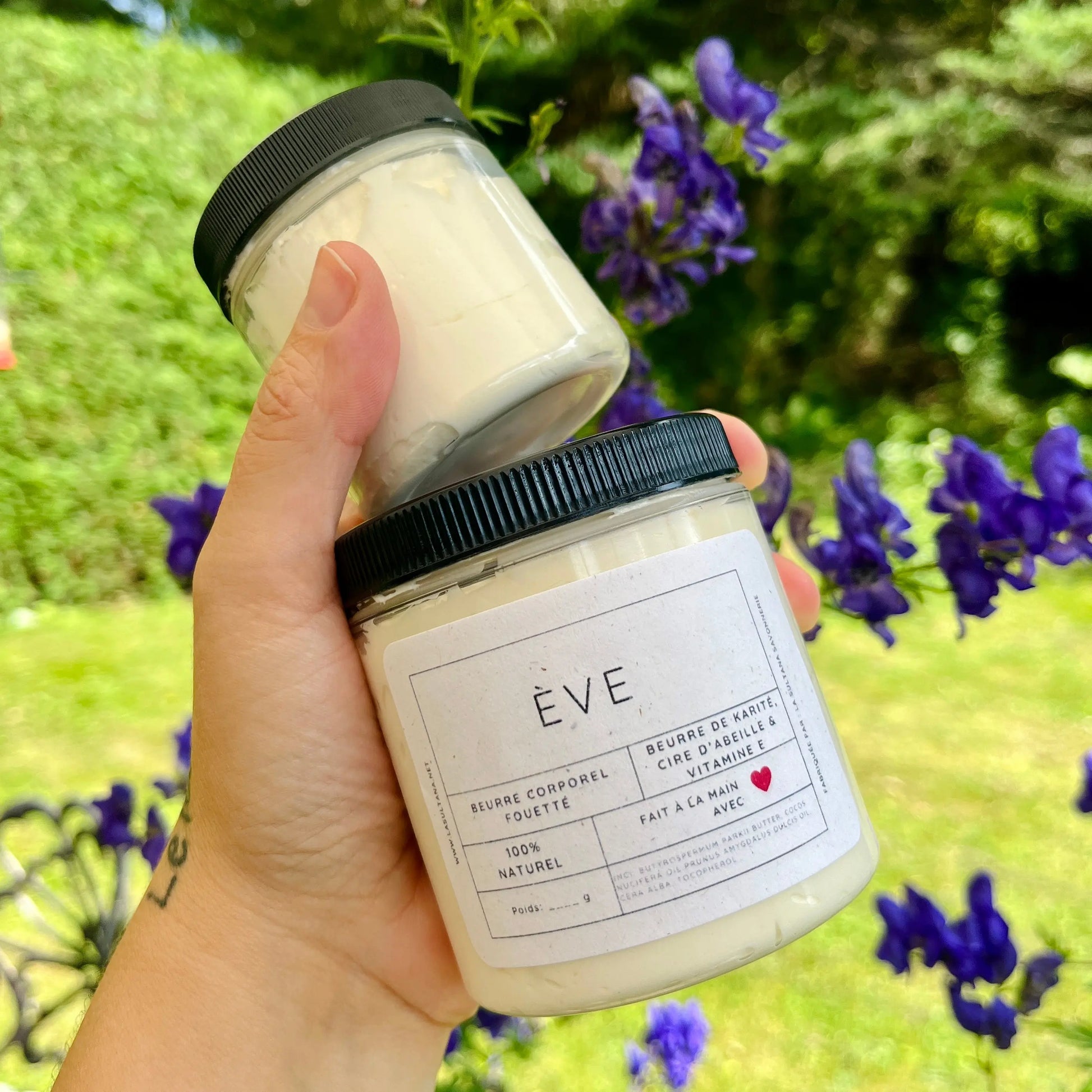 Une main tenant deux pots de « Beurre corporel fouetté ÈVE au karité » de La Sultana Savonnerie | Savons et soins naturels faits main | Québec. Le plus grand pot, avec une étiquette blanche comportant du texte et un petit cœur rouge, indique qu'il est 100 % naturel et fait de beurre de karité, de gousse de vanille et de vitamine E. Des fleurs violettes et de la verdure ornent l'arrière-plan.