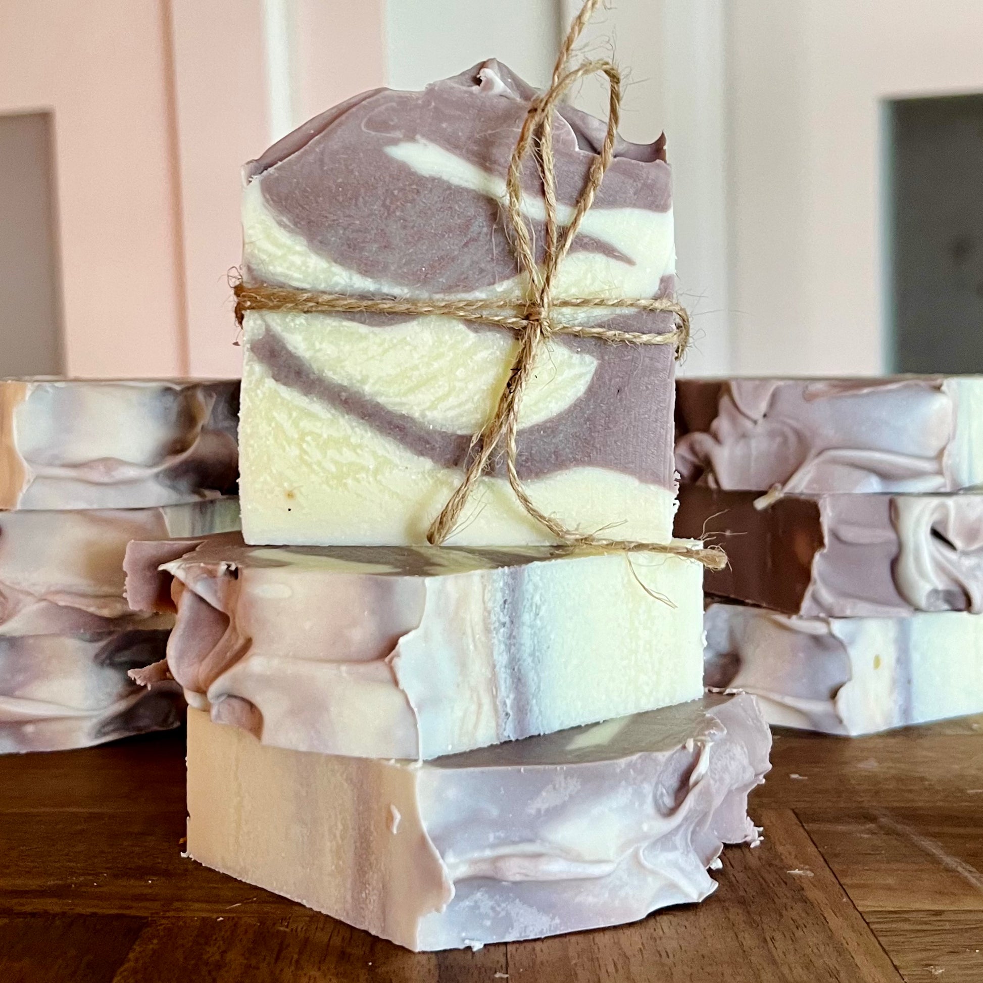 Trois morceaux de Savon à l'argile brésilienne WHITNEY de La Sultana Savonnerie, fabriqués avec de l'huile d'avocat et lilas, empilés sur une surface en bois. Le savon sur le dessus est attaché avec un morceau de ficelle. D’autres savons similaires sont en arrière-plan. Le savon présente un motif marbré dans des tons de violet et de blanc.