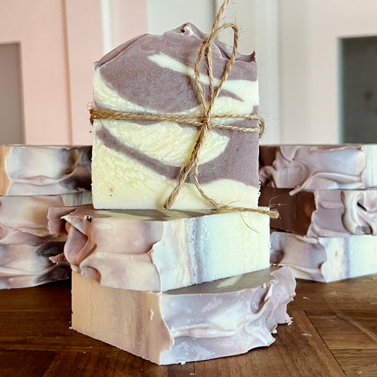 Trois morceaux de Savon à l'argile brésilienne WHITNEY de La Sultana Savonnerie, fabriqués avec de l'huile d'avocat et lilas, empilés sur une surface en bois. Le savon sur le dessus est attaché avec un morceau de ficelle. D’autres savons similaires sont en arrière-plan. Le savon présente un motif marbré dans des tons de violet et de blanc.