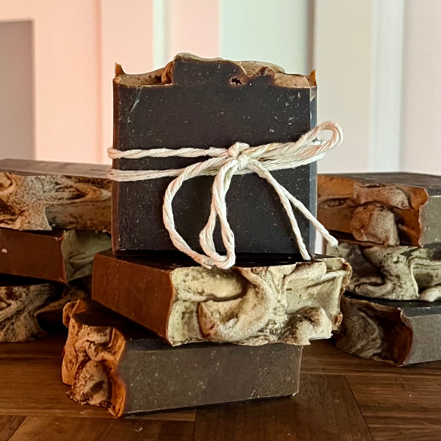 Une pile de Savon doux MICAELA beurre de cacao et chocolat de La Sultana Savonnerie | Savons et soins naturels faits main | Québec, avec une barre supérieure nouée avec une ficelle blanche. Les savons ont un aspect marbré multicolore avec des dessus texturés et sont enrichis en beurre de cacao pour des propriétés extra nourrissantes. Ils sont posés sur une surface en bois avec un arrière-plan flou.