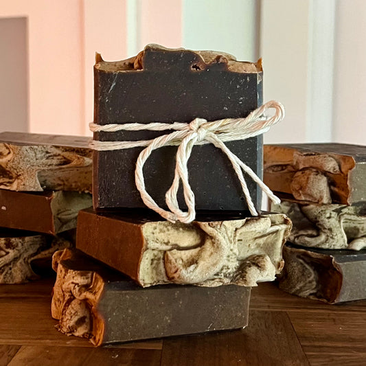 Une pile de Savon doux MICAELA beurre de cacao et chocolat de La Sultana Savonnerie | Savons et soins naturels faits main | Québec, avec une barre supérieure nouée avec une ficelle blanche. Les savons ont un aspect marbré multicolore avec des dessus texturés et sont enrichis en beurre de cacao pour des propriétés extra nourrissantes. Ils sont posés sur une surface en bois avec un arrière-plan flou.