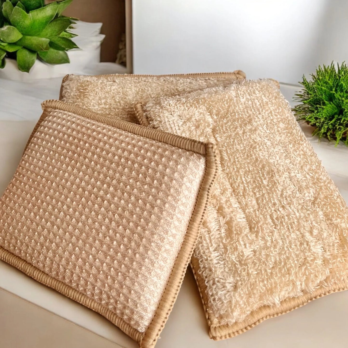 Trois serviettes beiges texturées sont soigneusement disposées sur une surface blanche. L'une d'elles a une texture gaufrée, tandis que les deux autres ont une texture moelleuse et bouclée. En arrière-plan, des plantes vertes et l'Éponge en bambou écologique de La Sultana Savonnerie | Savons et soins naturels faits main | Québec ajoutent une touche de couleur à la scène.