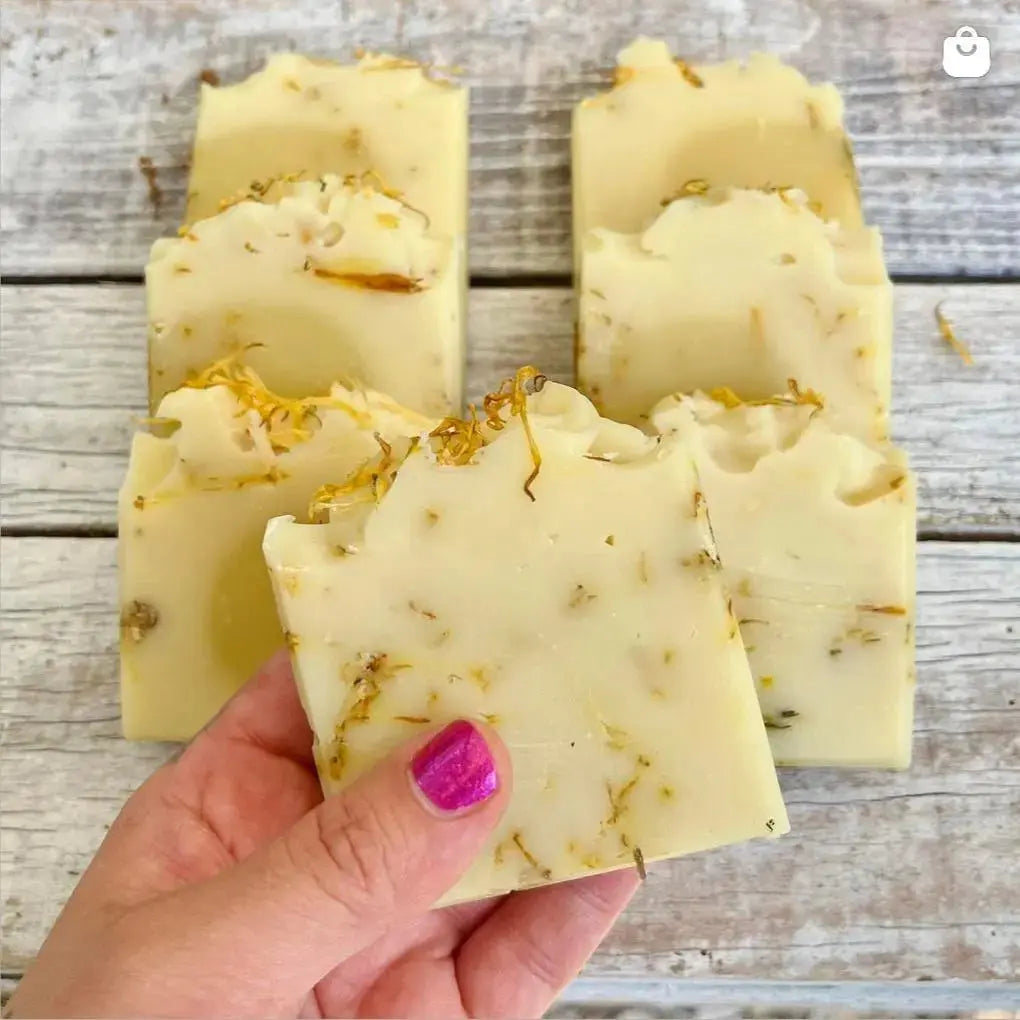 Une main au vernis à ongles rose tient une barre de Savon doux DAISY à l'aloe vera et calendula, à base d'ingrédients naturels, avec des herbes séchées visibles. En arrière-plan, quatre barres similaires de savon naturel sont disposées sur une surface en bois vieilli. Le pseudo Instagram @la_sultana_savonnerie est affiché en bas à gauche. La Sultane Savonnerie | Savons et soins naturels faits main | Québec.