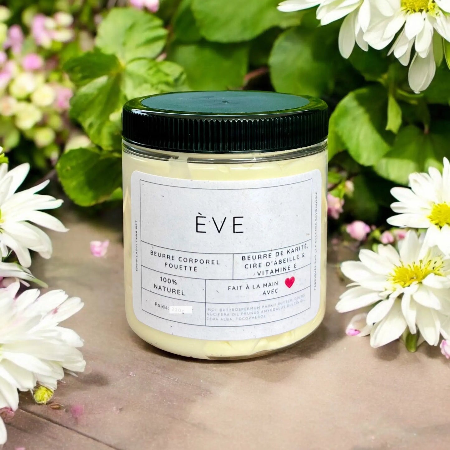 Un pot de beurre corporel fouetté ÈVE au karité de La Sultana Savonnerie est placé parmi des marguerites blanches et de la verdure. L'étiquette indique qu'il contient du beurre de karité, de la cire d'abeille et de la vitamine E, qu'il est fait à la main et 100 % naturel. Le pot blanc a un couvercle noir et un symbole de cœur à côté des mots « fait à la main ».
