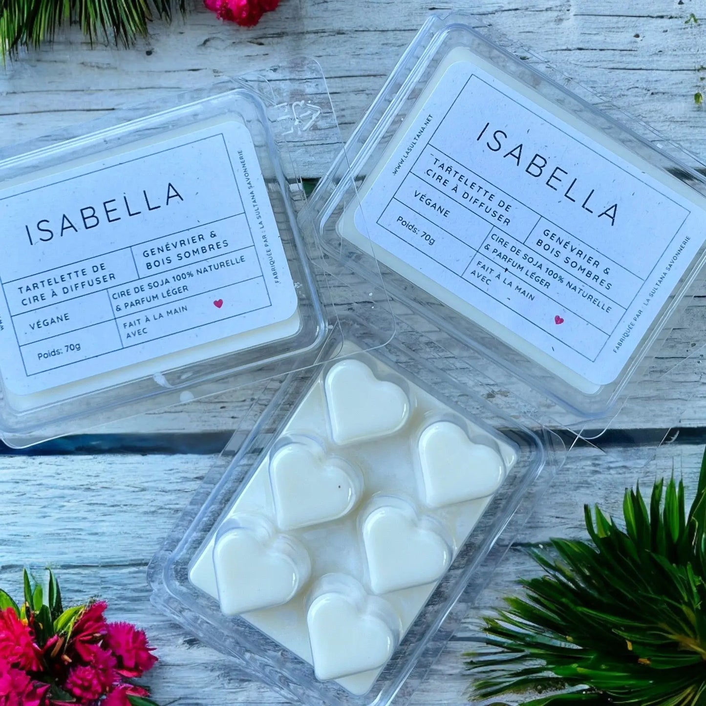 Deux emballages de Fondant parfumé au soja étiquetés « Tartelette au soja ISABELLA genévrier et bois sombres » de La Sultana Savonnerie | Savons et soins naturels faits main | Québec, contiennent des tartelettes à la cire de soja en forme de cœur. Un emballage est fermé et placé en angle, tandis que l'autre est ouvert, affichant six cœurs blancs. Autour des emballages se trouvent quelques fleurs rouges et des branches de pin vertes sur une surface en bois foncé.
