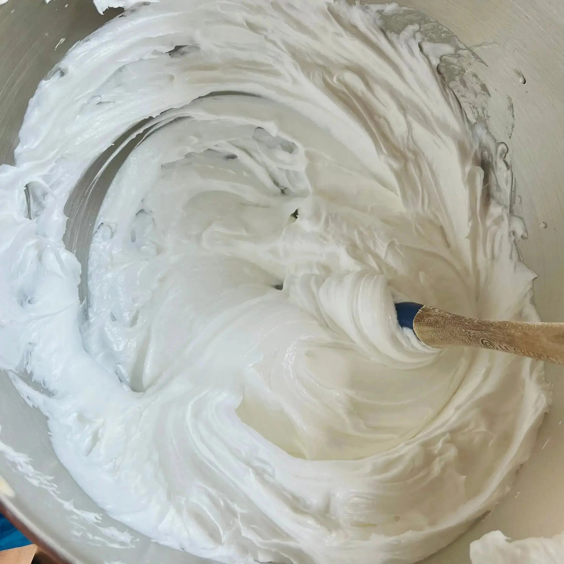 Un bol à mélanger contient un mélange succulent et épais qui est agité avec une cuillère en bois. La texture onctueuse et brillante, qui rappelle le savon fouetté FAIROUZ agrûmes sucrés de La Sultana Savonnerie | Savons et soins naturels faits main | Le Québec présente des motifs tourbillonnants à sa surface.