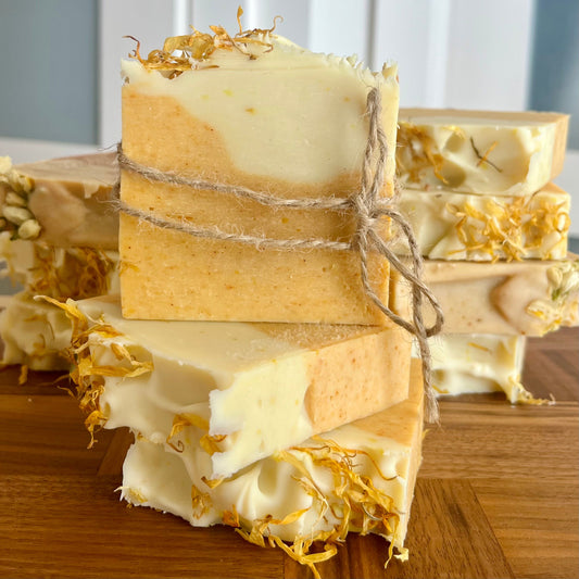 Une pile de savons doux  faits à la main à la cire d'abeille, au curcuma et à la camomille de La Sultana Savonnerie | Savons et soins naturels faits main | Québec est posée sur une surface en bois. Le savon présente un design bicolore avec un dessus crémeux et un dessous beige, orné de pétales de calendula séchés. Une barre est enveloppée de ficelle, sur fond de mur blanc.