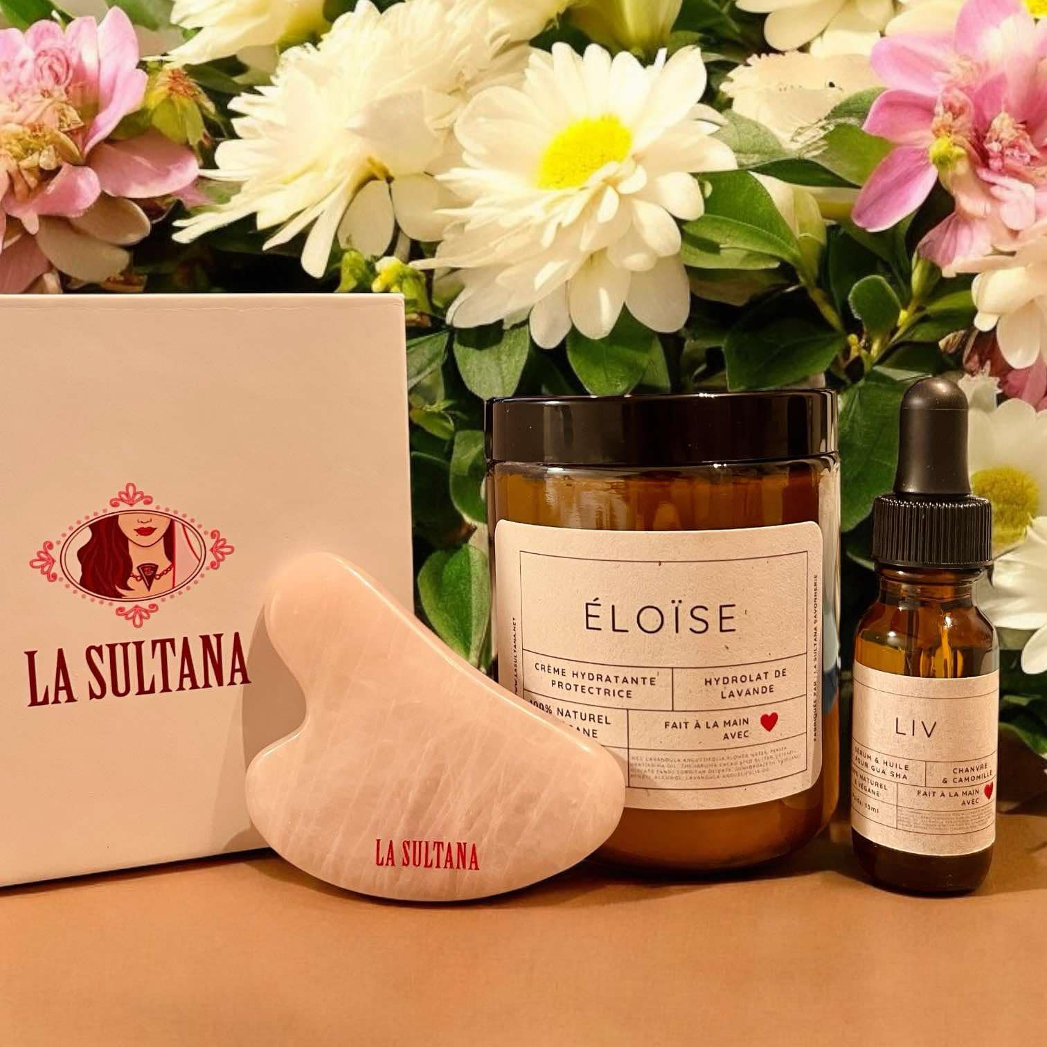 Une vitrine de produits de soins de la peau Éloïse et Liv présente un pot de crème hydratante et un flacon compte-gouttes de sérum de chanvre, accompagnés de l'outil de massage en quartz rose Black Friday JOUR 2 de La Sultana Savonnerie. L'arrangement est placé sur un fond de fleurs roses et blanches, avec la boîte de La Sultana Savonnerie visible à gauche.