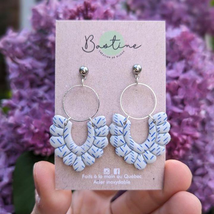 Une main tient une paire de Boucles d'oreilles PÉNÉLOPE sur fond de fleurs violettes. Les boucles d'oreilles comportent des anneaux en argent avec des pendants de pièces à motifs blancs et bleus. L'étiquette indique « La Sultana Savonnerie, création de bijoux. Faits à la main au Québec. Hypoallergènes, acier inoxydable.