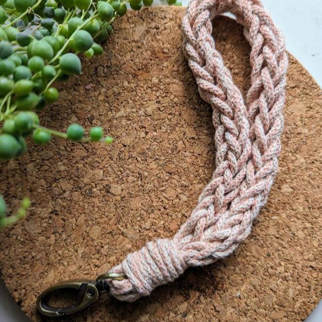 Un Porte-clé fait au crochet de La Sultana Savonnerie | Savons et soins naturels faits main | Québec, avec un clip en métal, repose sur une surface ronde en liège à côté d'une plante collier de perles. Fabriqué par un artisan de Québec, il allie élégance fonctionnalité et esthétique.