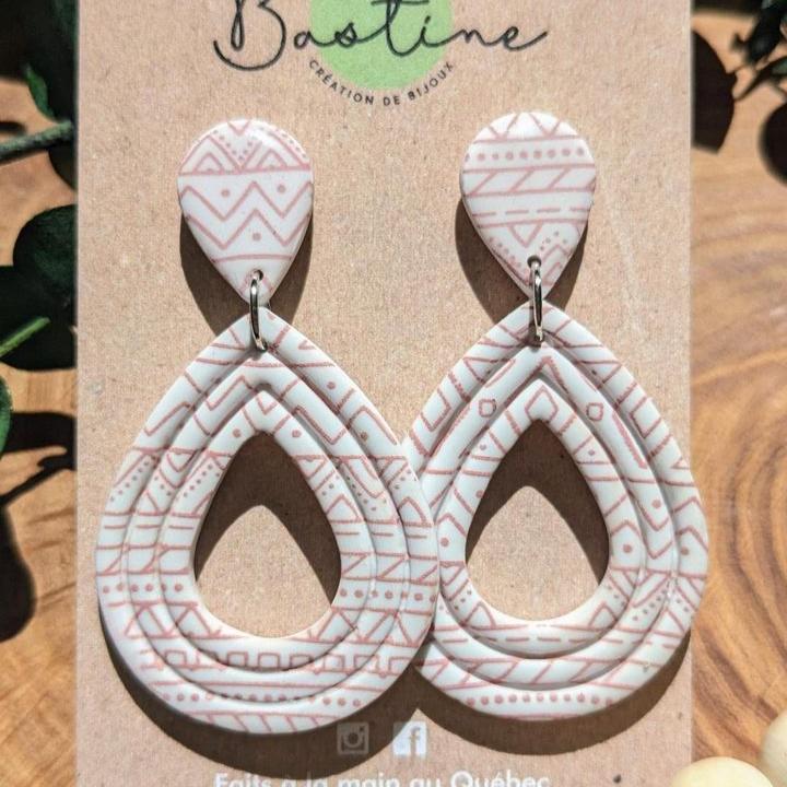 Une paire de boucles d'oreilles pendantes hypoallergéniques en forme de goutte, appelées Boucles d'oreilles CORALIE, arborent des motifs géométriques roses et blanc cassé. Présentées sur une carte portant le nom de marque « La Sultana Savonnerie » et « Savons et soins naturels faits main | Québec », elles mettent fièrement en valeur leur origine artisanale au Québec.