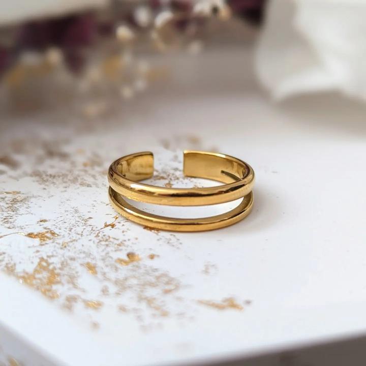 Une bague ouverte en or, la « Bague double plaquée 18k », au design minimaliste à double ligne, est posée sur une surface blanche ornée de taches d'or éparses. Fabriquée par la Savonnerie La Sultana de Québec, l'allure sophistiquée de la bague est complétée par des éléments flous de fleurs séchées et un objet blanc en arrière-plan.