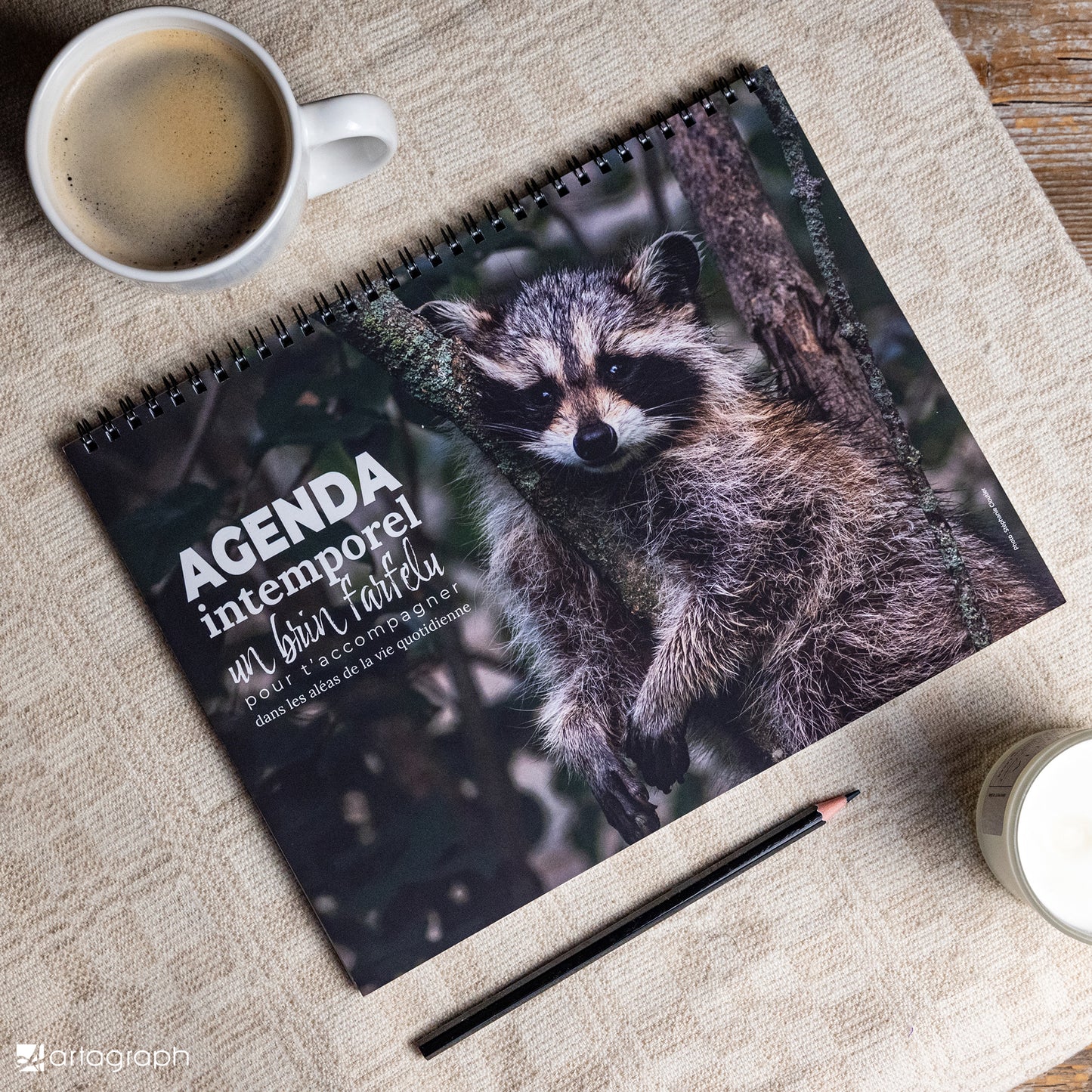 Sur une table en bois repose un calendrier à reliure spirale sur lequel figure un raton laveur. Il est accompagné d'une tasse de café, d'une bougie allumée et d'un crayon qui donne le ton. Le texte « Agenda intemporel RATON-LAVEUR » capture parfaitement l'essence intemporelle d'un agenda élégant et non daté. Ce produit est offert par La Sultana Savonnerie | Savons et soins naturels faits main | Québec.