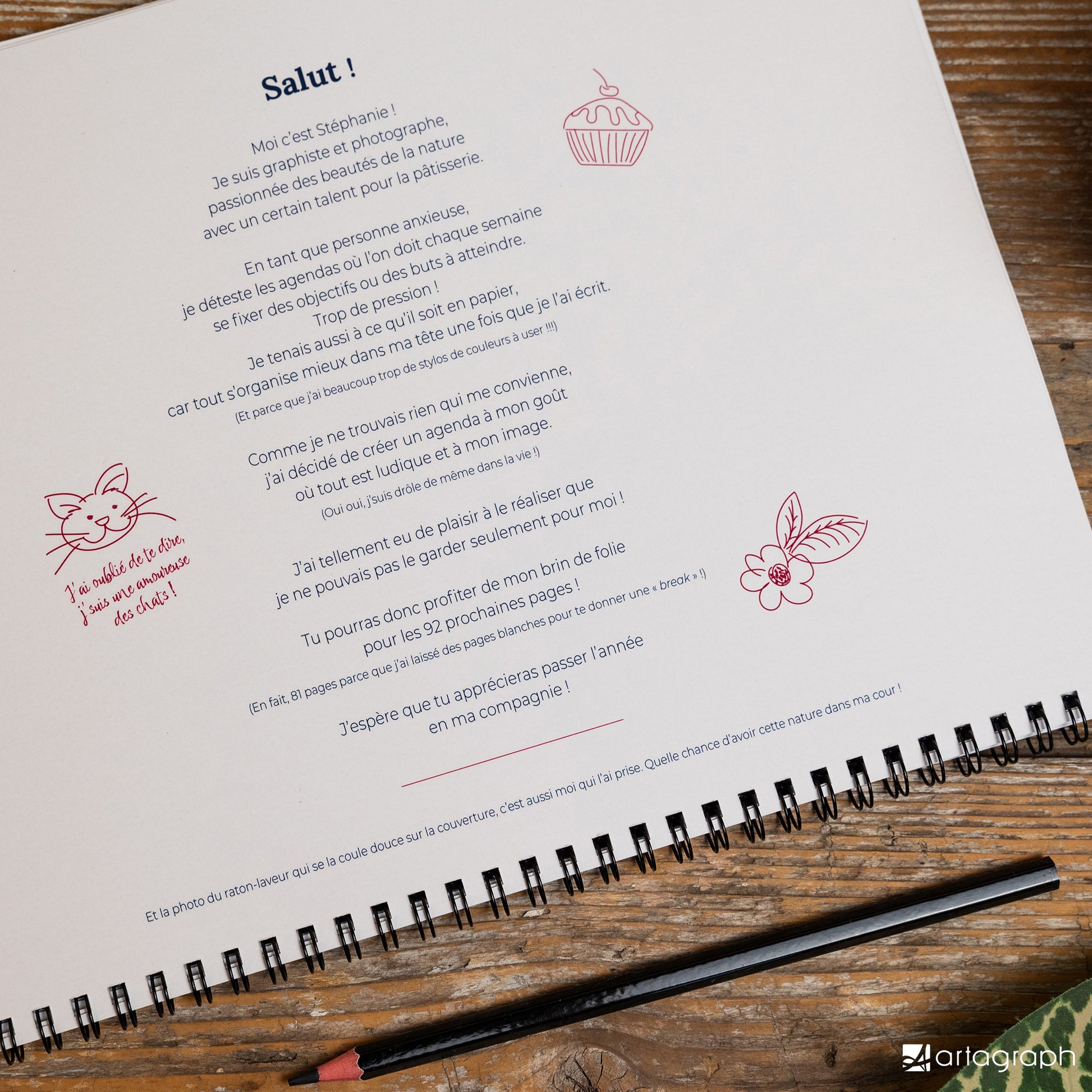 Une page de carnet ouverte présente un texte français manuscrit aux côtés de petits gribouillis représentant un petit gâteau, un chat et une tasse. Posé sur une surface en bois avec un trombone sur le bord, cet Agenda intemporel RATON-LAVEUR de La Sultana Savonnerie, fabricant de savons et de soins naturels artisanaux au Québec, est doté d'une couverture « artographe », idéale pour tout planificateur créatif en quête de style.
