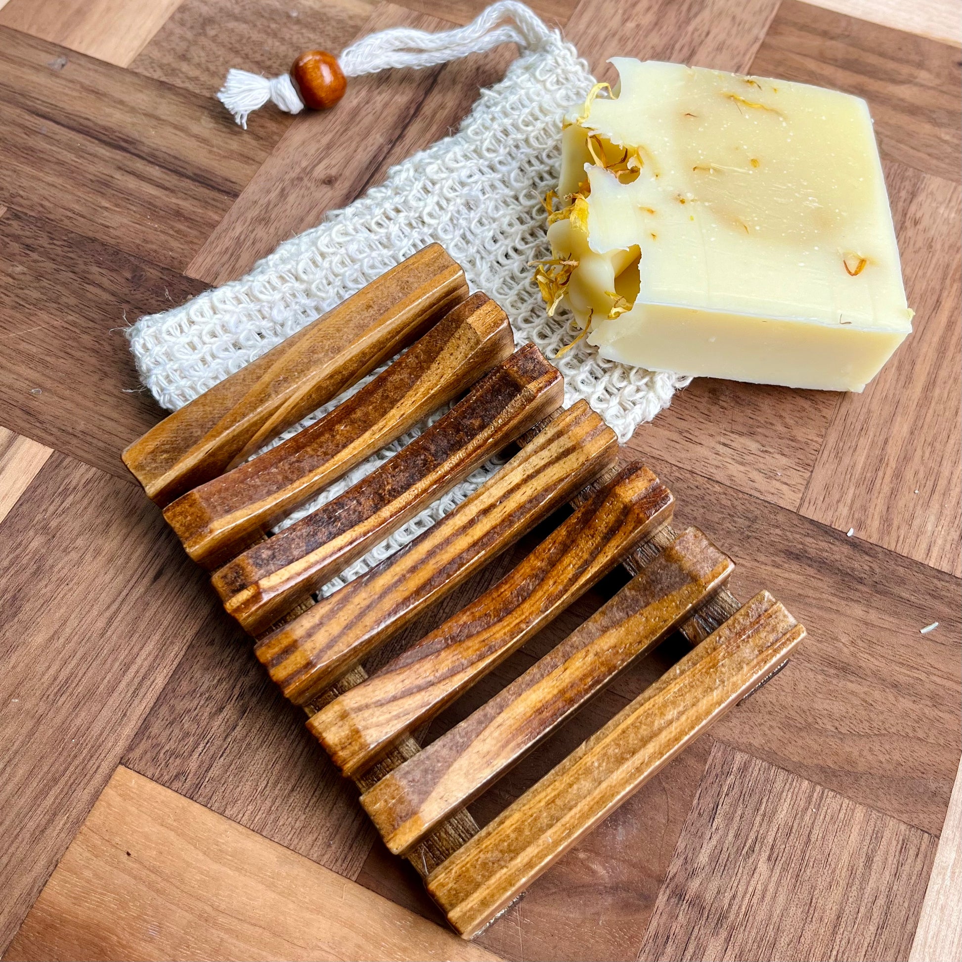 Une barre de savon artisanal aux pétales de fleurs séchées repose sur un sac-économiseur de savon en sisal blanc de La Sultana Savonnerie | Savons et soins naturels faits main | Québec. À côté d'eux se trouve un porte-savon en bois de pin à lattes horizontales, le tout posé sur une surface en bois à motif en damier.