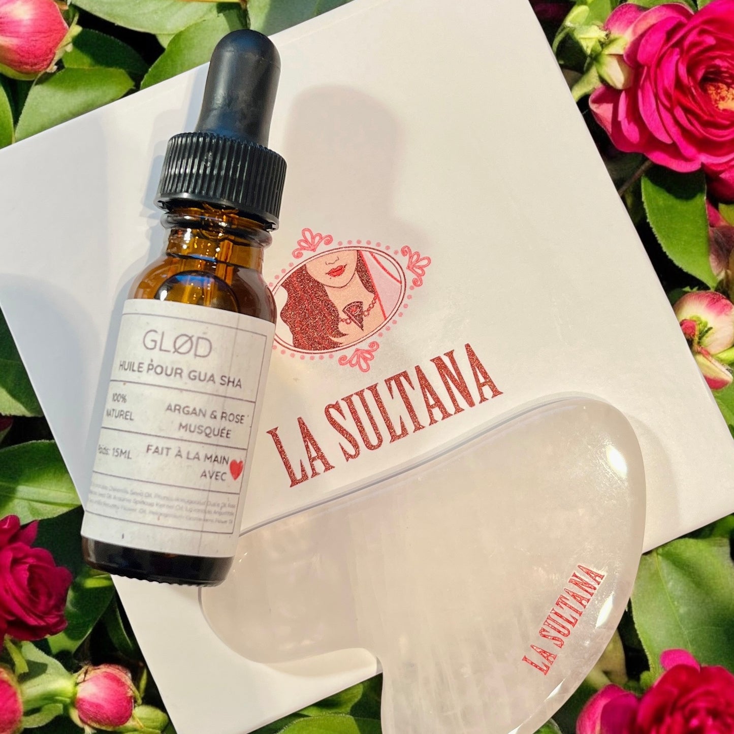 Un flacon d'huile pour le visage Glød avec compte-gouttes, étiqueté « Huile pour Gua Sha », infusé d'huiles d'argan et de rose, est élégamment placé dans un emballage « La Sultana Savonnerie ». À côté, l'exquis outil Gua Sha en quartz rose se trouve au milieu de roses rouges, offrant une expérience de soin de la peau luxueuse.