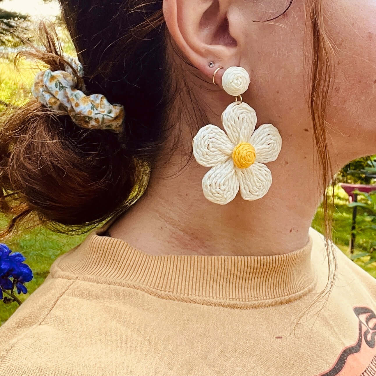 Un personnage aux cheveux roux attachés par un chouchou de la collection "Chouchous à cheveux faits à partir de retailles (paquet de 3)" de La Sultana Savonnerie est représenté de profil, portant une grande boucle d'oreille blanche et jaune en forme de fleur. Le personnage est vêtu d'un haut beige et se tient debout dans un décor extérieur avec de l'herbe verte et quelques fleurs visibles.