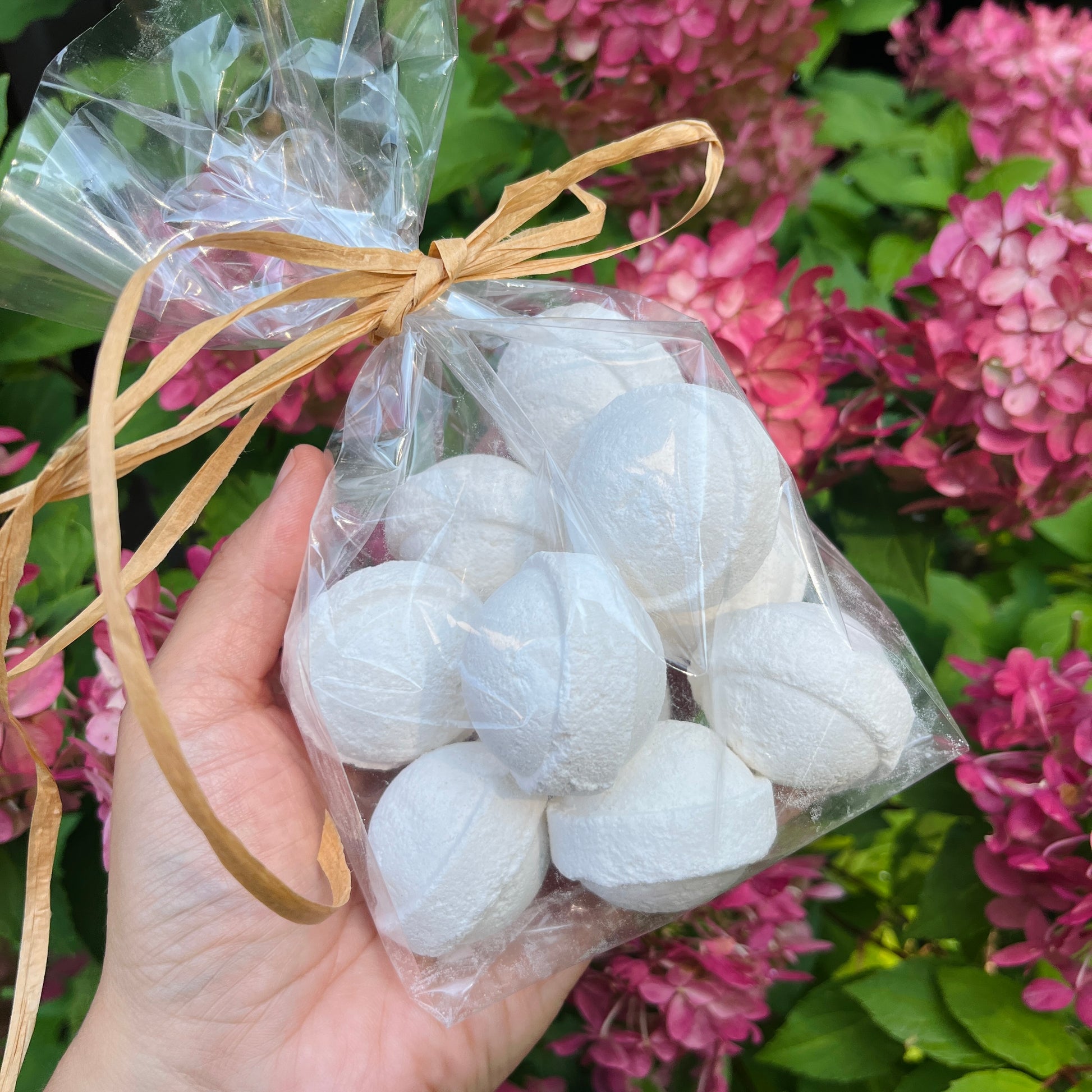 Une main tient un sac en plastique transparent rempli de bombes de bain rondes EMILY parfumées à la lavande, soigneusement attachées avec une ficelle rustique. Le fond met en valeur des fleurs d'hortensia roses en fleurs et un feuillage vert luxuriant, mettant en valeur la beauté naturelle. Ces bombes de bain sont fabriquées par La Sultana Savonnerie | Savons et soins naturels faits main | Québec.