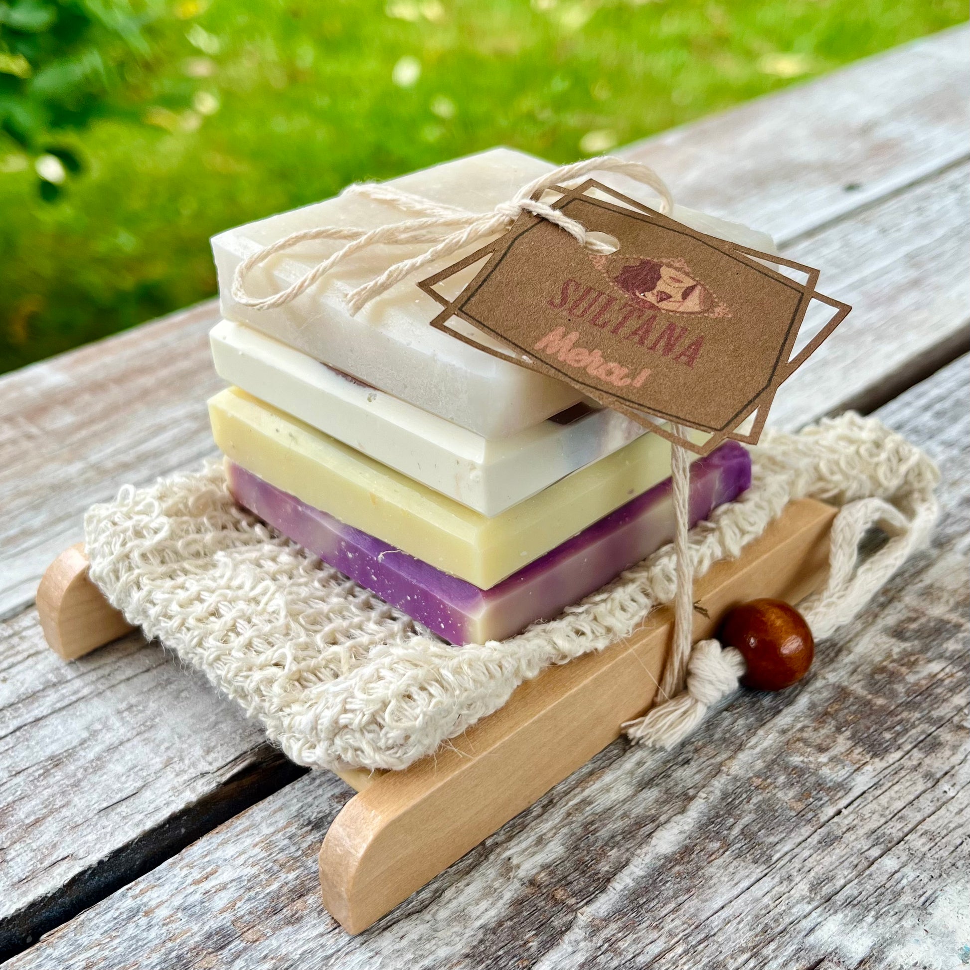 Une pile de mini savons naturels multicolores est nouée avec de la ficelle et placée sur un petit porte-savon en bois muni d'une brosse beige. L'Ensemble MIGNONNE porte une étiquette marron intitulée « Suzana Merci ». L'arrière-plan présente une zone herbeuse et une surface en bois patinée, complétant l'ambiance artisanale de La Sultana Savonnerie | Savons et soins naturels faits main | Québec.