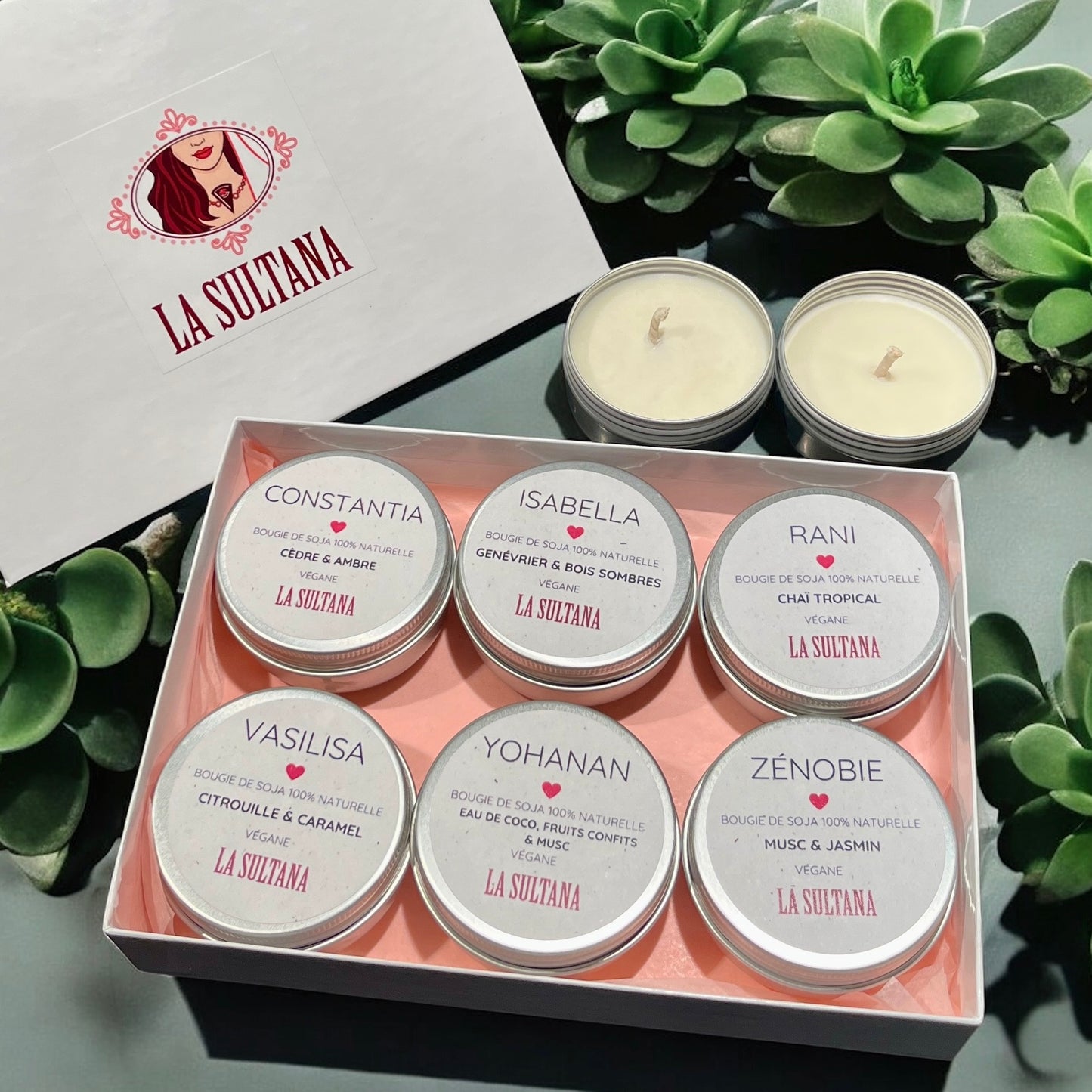 Un coffret de présentation contient l'ensemble « LUZ », composé de six bougies parfumées La Sultana Savonnerie fabriquées à partir de cire de soja. Chaque bougie est logée dans une boîte ronde, chacune étiquetée avec des noms et des parfums distinctifs : Constantia, Isabella, Rani, Vasilisa, Yohanan et Zenobie. Le couvercle met en valeur le nom de la marque « La Sultana Savonnerie | Savons et soins naturels faits main | Québec », tandis que de petites plantes entourent naturellement cet élégant emballage.