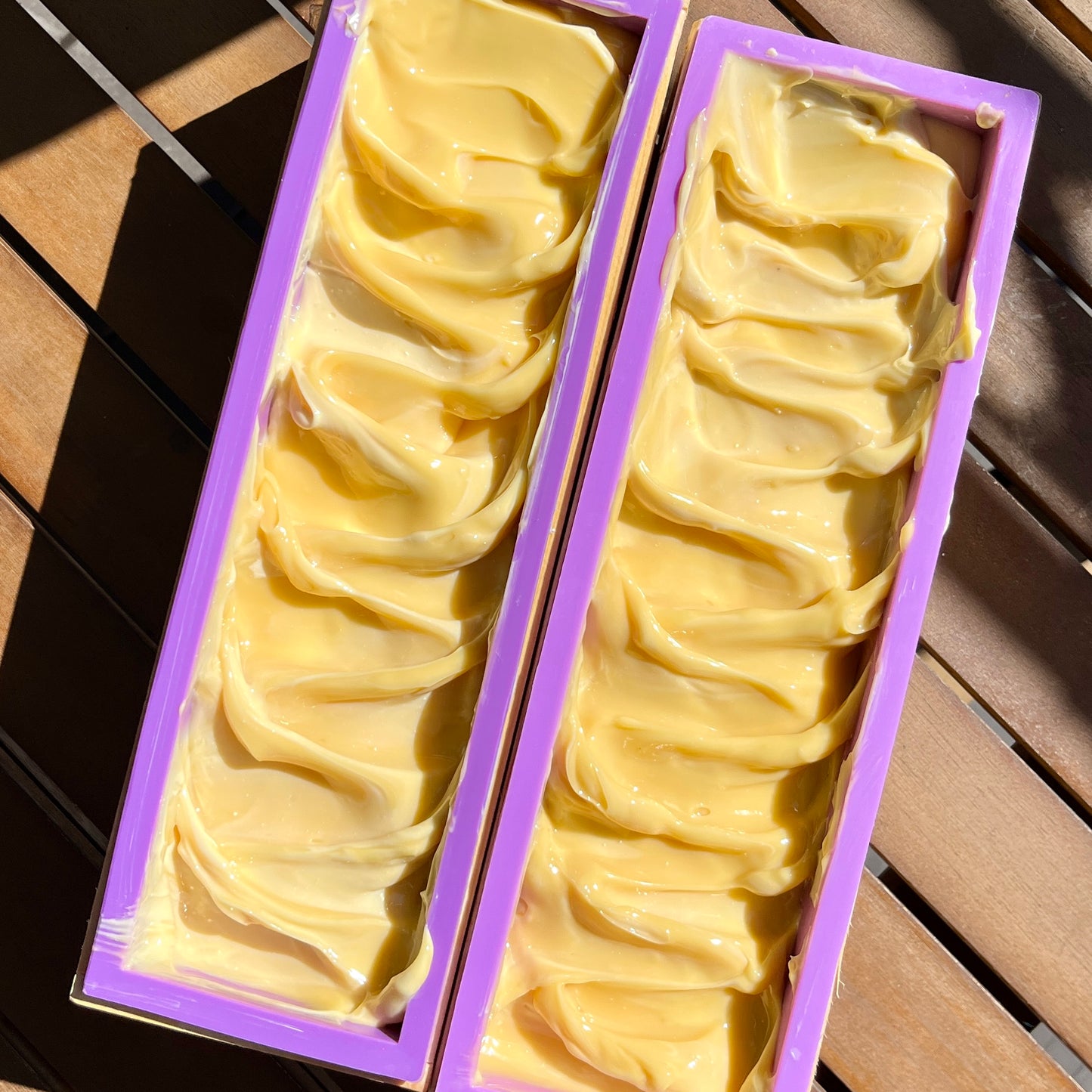 Deux moules rectangulaires de Savon doux RANAVALONA vanille à la crème, remplis de pâte à savon jaune crémeuse enrichie de beurre de cacao, sont placés sur une surface en bois. Le savon artisanal de La Sultana Savonnerie | Savons et soins naturels faits main | Québec a une texture lisse avec un motif ondulé sur le dessus. Chaque moule est bordé d'une bordure violette.