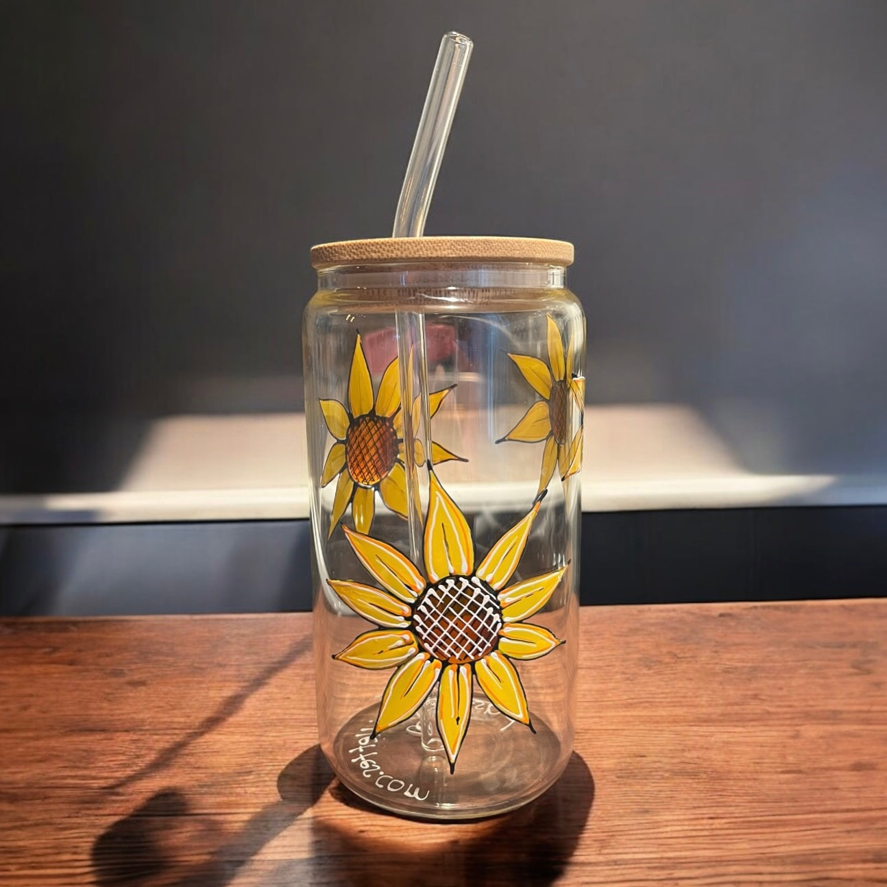 Un verre peint à la main Tournesol, fabriqué par La Sultana Savonnerie | Savons et soins naturels faits main | Québec, est présenté sur une surface en bois. Ce verre transparent est accompagné de tournesols jaunes peints à la main, d'un couvercle en bambou et d'une paille transparente. Le fond reflète l'inspiration québécoise avec son mur gris à motifs d'ombre et de lumière.