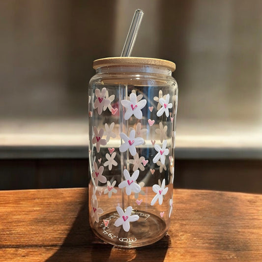 Un pot en verre transparent de la collection Verres peints à la main Marguerites & Coeurs de La Sultana Savonnerie, doté d'un couvercle et d'une paille en bambou, est orné de fleurs blanches et roses en forme de cœur, élégamment placées sur une surface en bois sur un fond flou.