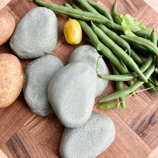 Une surface en bois présente plusieurs pierres grises lisses, quelques pommes de terre, une tomate jaune, une botte de haricots verts frais et un Savon exfoliant INARI aloe vera & pierre ponce de La Sultana Savonnerie | Savons et soins naturels faits main | Québec.