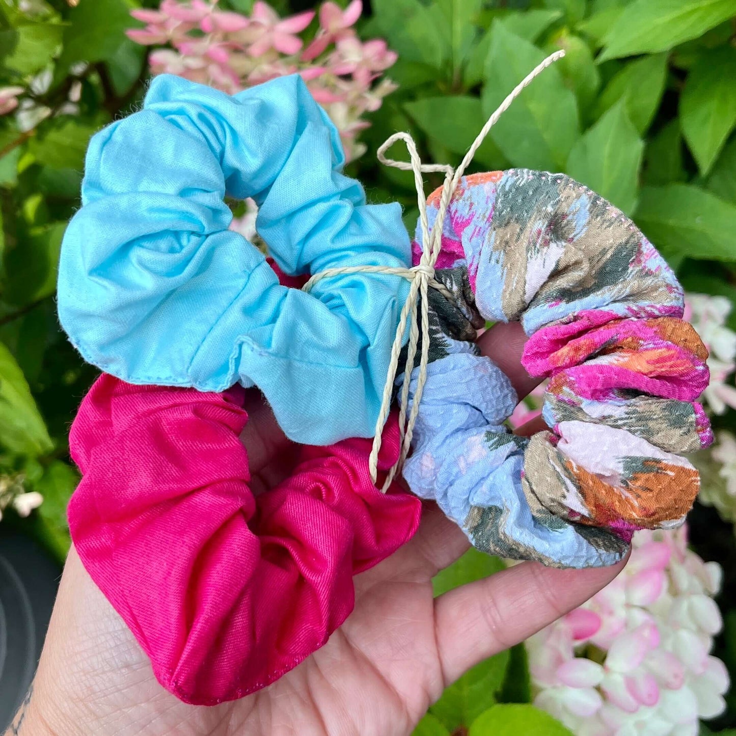 Une main tient trois chouchous à cheveux colorés faits à partir de la collection artisanale de La Sultana Savonnerie : un bleu, un rose et un multicolore avec un petit noeud en ficelle. Le fond est composé de feuilles vertes et de fleurs roses, mettant en valeur ces magnifiques accessoires fabriqués au Québec.