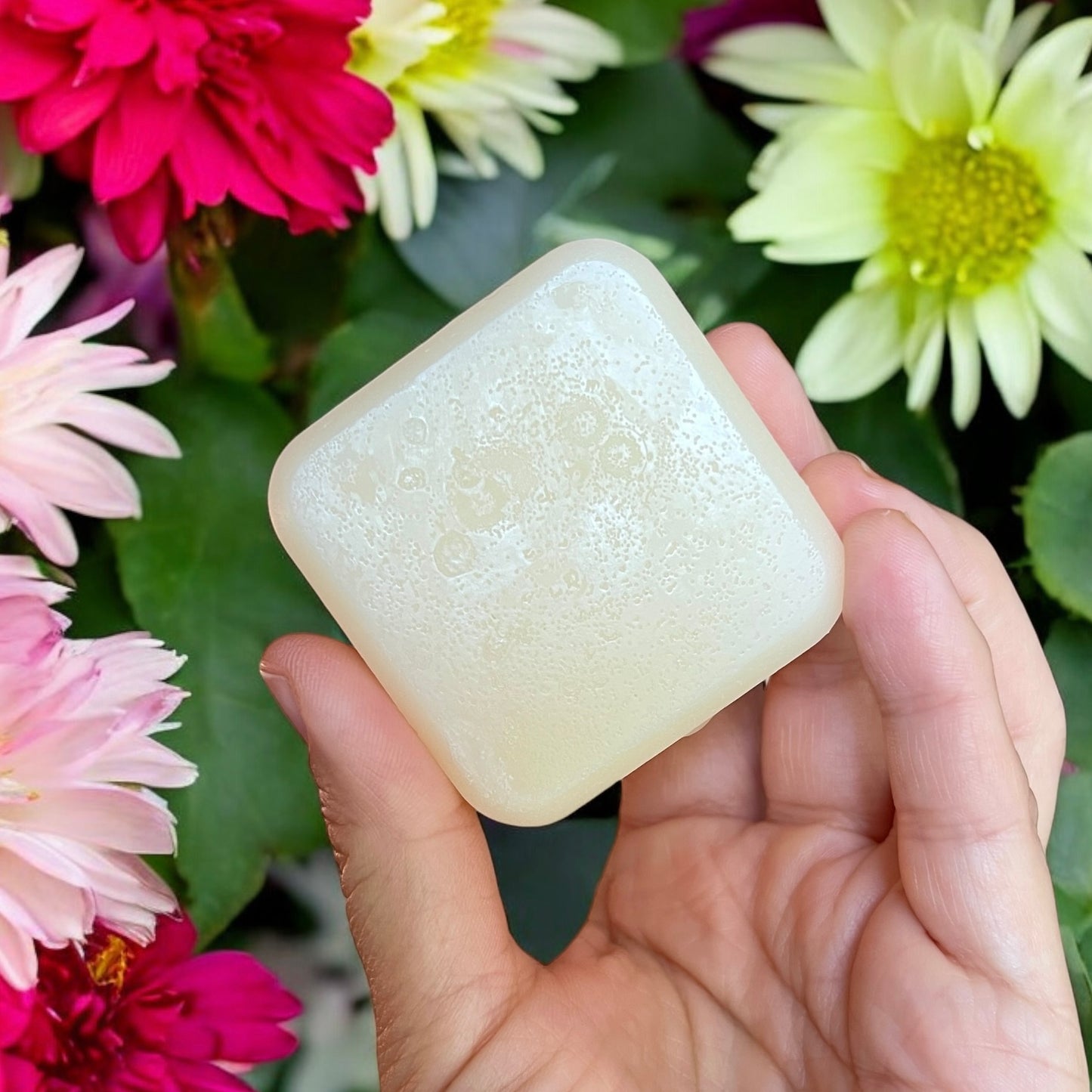 Une main présente une barre carrée de savon « Revitalisant naturel YEVA beurre de mangue et provitamine B5 » de La Sultana Savonnerie sur fond de fleurs éclatantes, mettant en valeur des fleurs roses, rouges et blanches avec des feuilles vertes luxuriantes. Le savon présente un aspect crémeux avec une surface légèrement texturée.