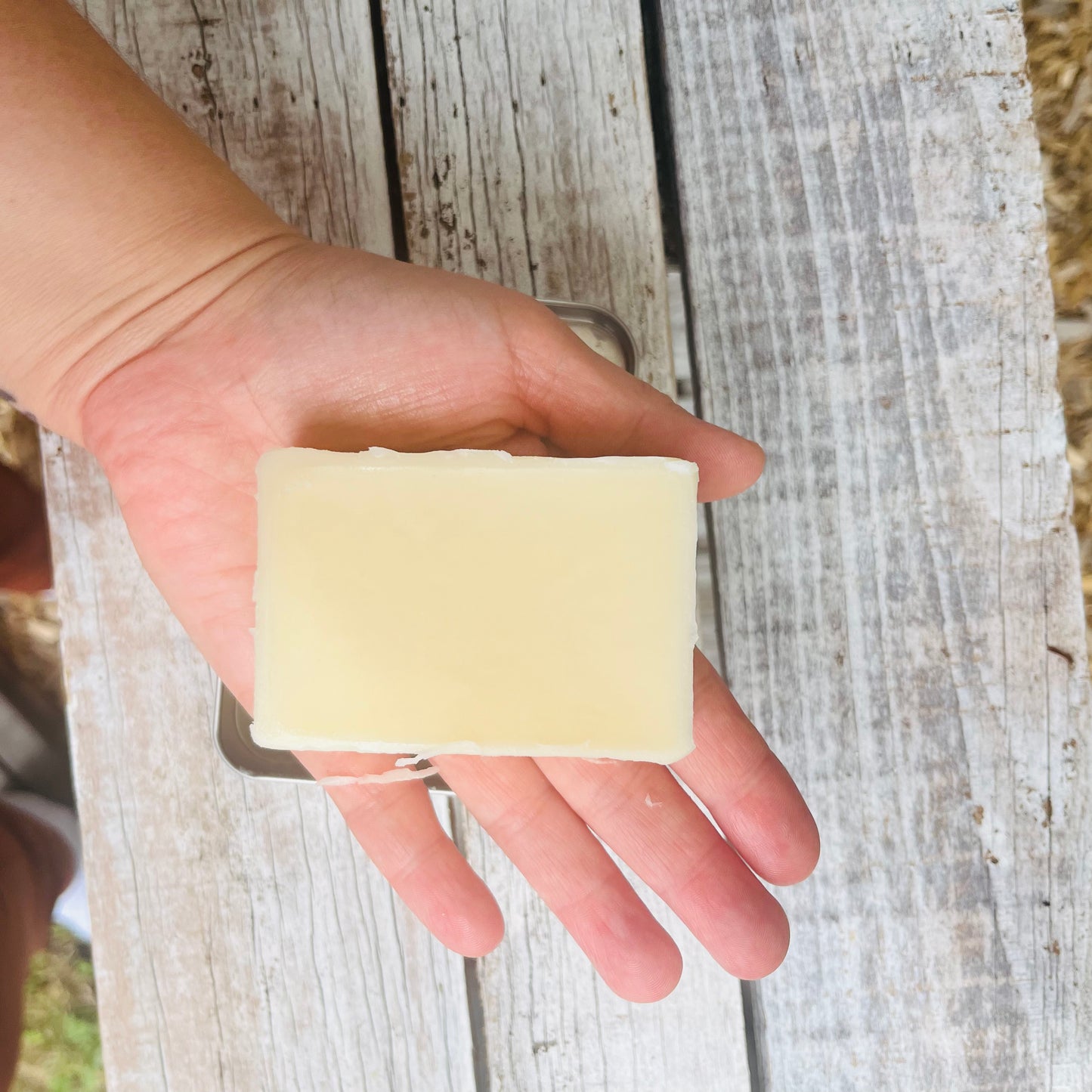 Une main tenant un pain de savon rectangulaire de couleur claire enrichi en beurre de karité KATE et en lotion à la vitamine E. Le fond est constitué de planches de bois patinées.