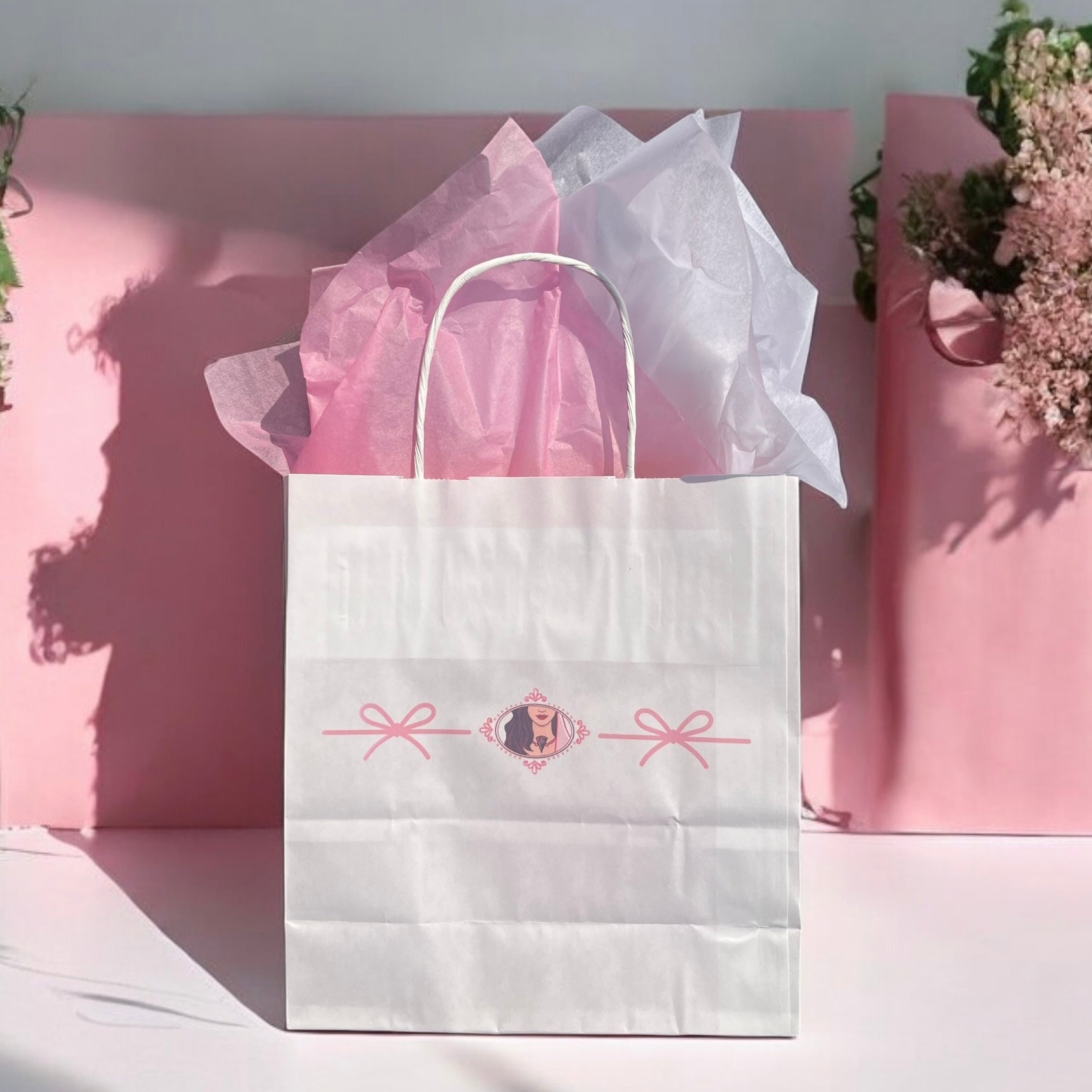 Un emballage cadeau blanc de Wrapped, rempli de papier de soie rose et blanc, est posé sur un fond rose. Cet élégant beau sac cadeau présente un charmant design de ruban rose et un petit emblème décoratif au centre. Positionnés sur le côté droit se trouvent de magnifiques compositions florales.