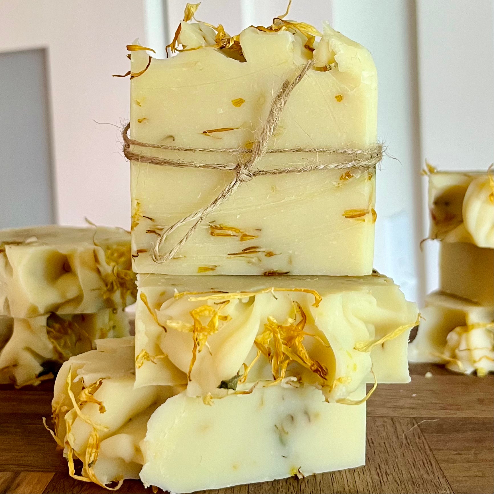 Une pile de Savon doux DAISY à l'aloe vera et calendula, infusé d'ingrédients hydratants, est nouée avec de la ficelle. Le savon naturel de La Sultana Savonnerie | Savons et soins naturels faits main | Le Québec est de couleur crème clair avec de petits morceaux de fleurs séchées incrustés dedans. Le savon semble être exposé sur une surface en bois à l'intérieur, parfait pour les peaux sensibles.