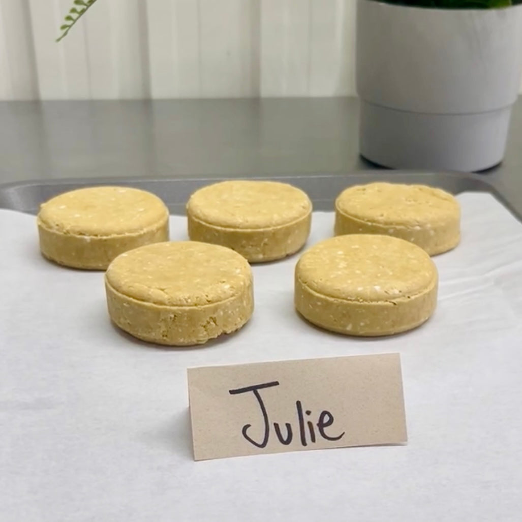 Une plaque à pâtisserie recouverte de papier sulfurisé contient cinq morceaux de pâte non cuite. Au premier plan, une petite pancarte affiche le nom manuscrit « Julie », rappelant le Shampoing naturel JULIE coco et écorce d'orange de La Sultana Savonnerie. Une plante en pot ornée de fleurs d'ylang ylang est partiellement visible à l'arrière-plan, ajoutant une touche de beauté naturelle.