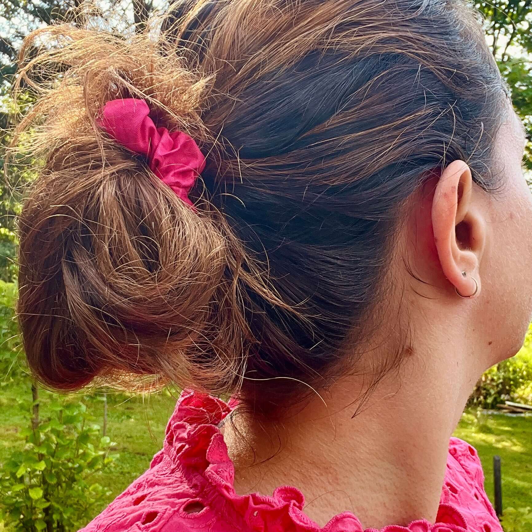 Une personne aux cheveux bruns attachés en chignon désordonné à l'aide d'un chouchou rose provenant des « Chouchous à cheveux faits à partir de retailles (paquet de 3) » de La Sultana Savonnerie | Savons et soins naturels faits main | Québec est représentée de profil et de dos. Elle porte un haut rouge à volants et se tient debout à l'extérieur avec de la verdure en arrière-plan.