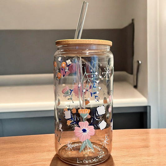 Une tasse en verre avec couvercle en bambou et paille transparente repose sur une table en bois, ornée de motifs floraux vibrants rappelant les « Verres peints à la main Tempête Florale ». En arrière-plan, un mur et une étagère aux couleurs douces et floues rehaussent cette scène enchanteresse de la collection québécoise La Sultana Savonnerie | Savons et soins naturels faits main.