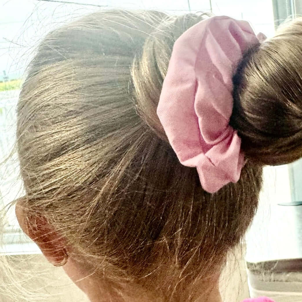 L'image en gros plan montre l'arrière de la tête d'une personne aux cheveux châtain clair attachés en chignon, maintenus par un chouchou rose à cheveux fait à partir de retailles. L'arrière-plan semble être un décor intérieur avec de grandes fenêtres. Ce chouchou fait partie du paquet de 3 de La Sultana Savonnerie | Savons et soins naturels faits main | Québec.