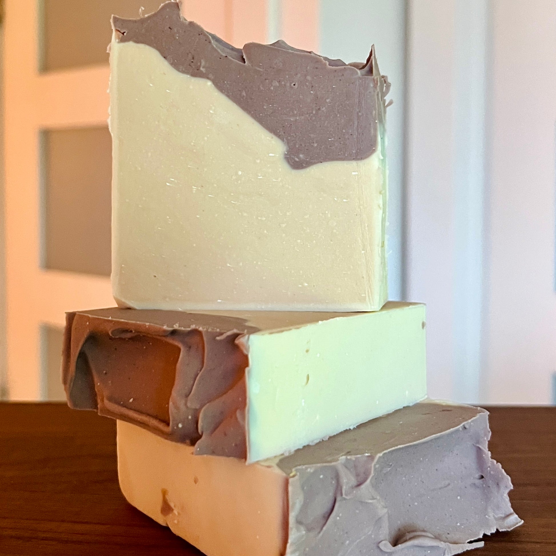 Trois barres de Savon au lait de coco GRACE avec argile brésilienne de La Sultana Savonnerie sont empilées sur une surface en bois. Chaque barre de savon a une base blanc crème avec des teintes brunâtres et grisâtres tourbillonnantes sur le dessus, idéales pour un usage quotidien. L'arrière-plan est légèrement flou, montrant une partie d'une porte et d'un mur, mettant en valeur la beauté artisanale de ces savons naturels faits à la main au Québec.