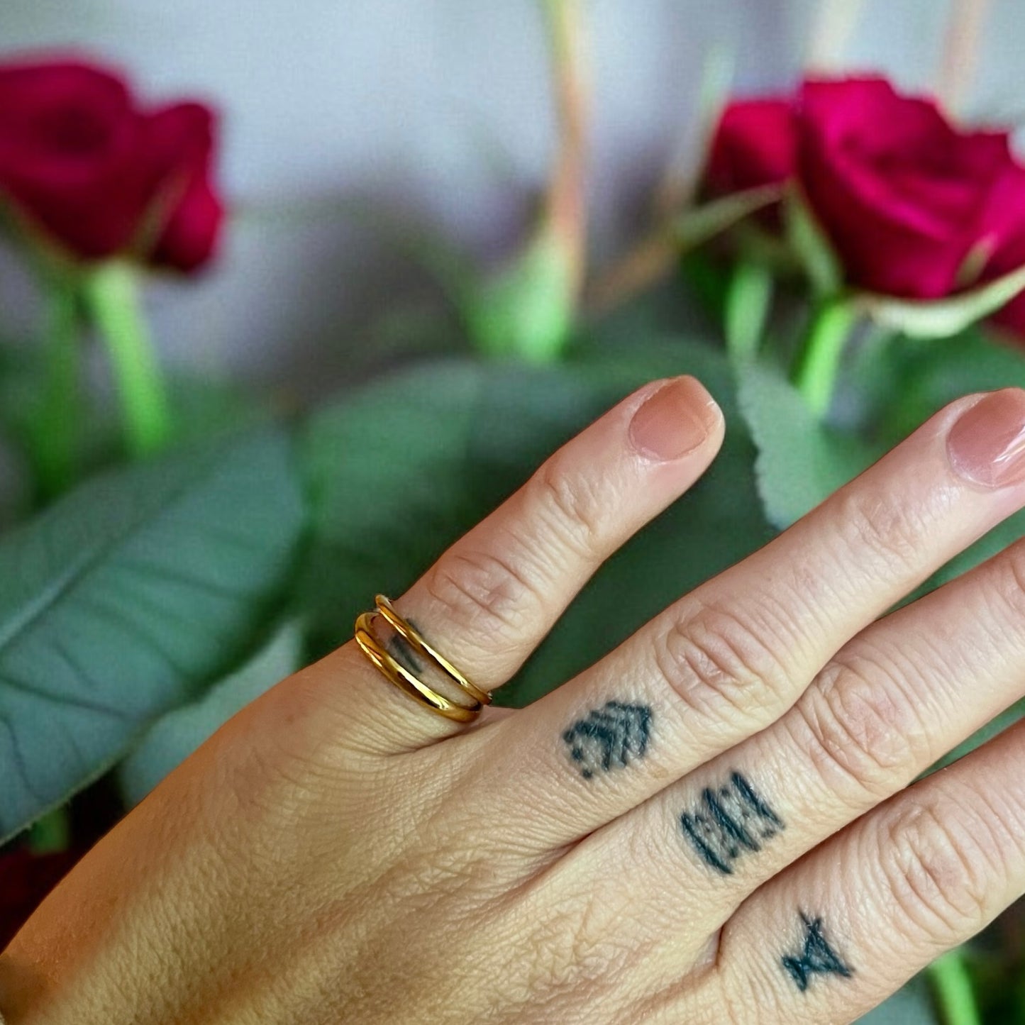 Une main ornée de deux tatouages arbore une Bague double plaquée 18k sur l'annulaire. En arrière-plan, des roses rouges éclatantes fleurissent sur des feuilles et des tiges d'un vert luxuriant. Le bijou fait partie de la collection La Sultana Savonnerie | Savons et soins naturels faits main | Québec.