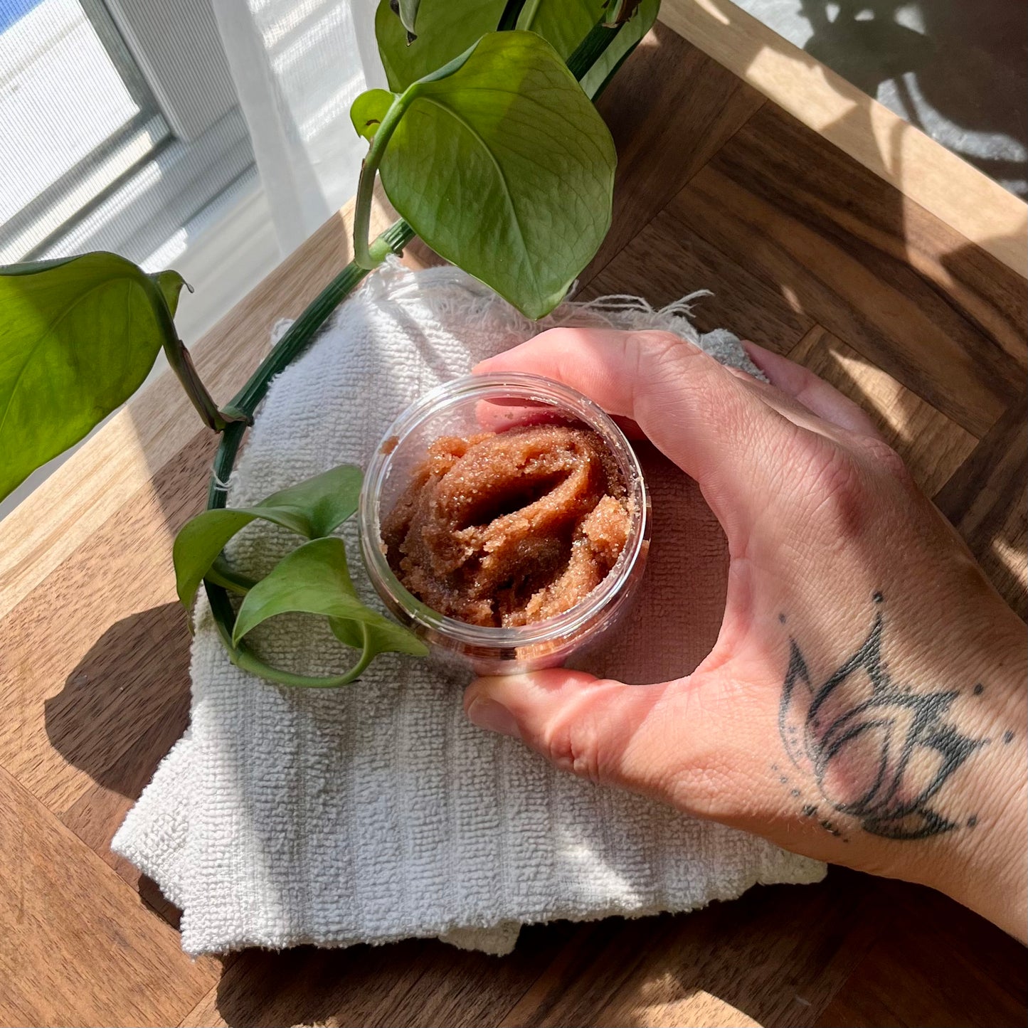 Une main tatouée tient un pot ouvert d'Exfoliant corporel STÉPHANIE chocolat et vanille de La Sultana Savonnerie sur une serviette blanche. La surface en bois et une plante d'intérieur verte sont visibles en arrière-plan. La lumière du soleil projette une ombre douce sur la scène, ajoutant à l’ambiance sereine tandis que vous régénérez votre peau grâce à ses éléments hydratants.