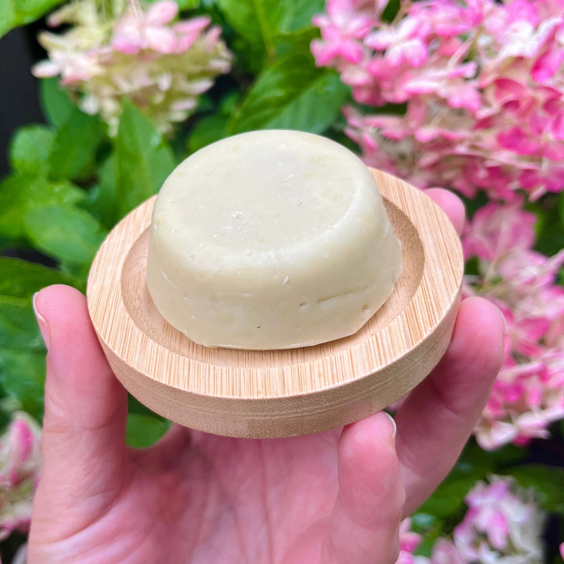 Une main tient un savon rond beige Crème à raser TIM romarin et chanvre de La Sultana Savonnerie sur un petit plat en bois. En arrière-plan, on distingue des feuilles vertes et des grappes de fleurs roses.