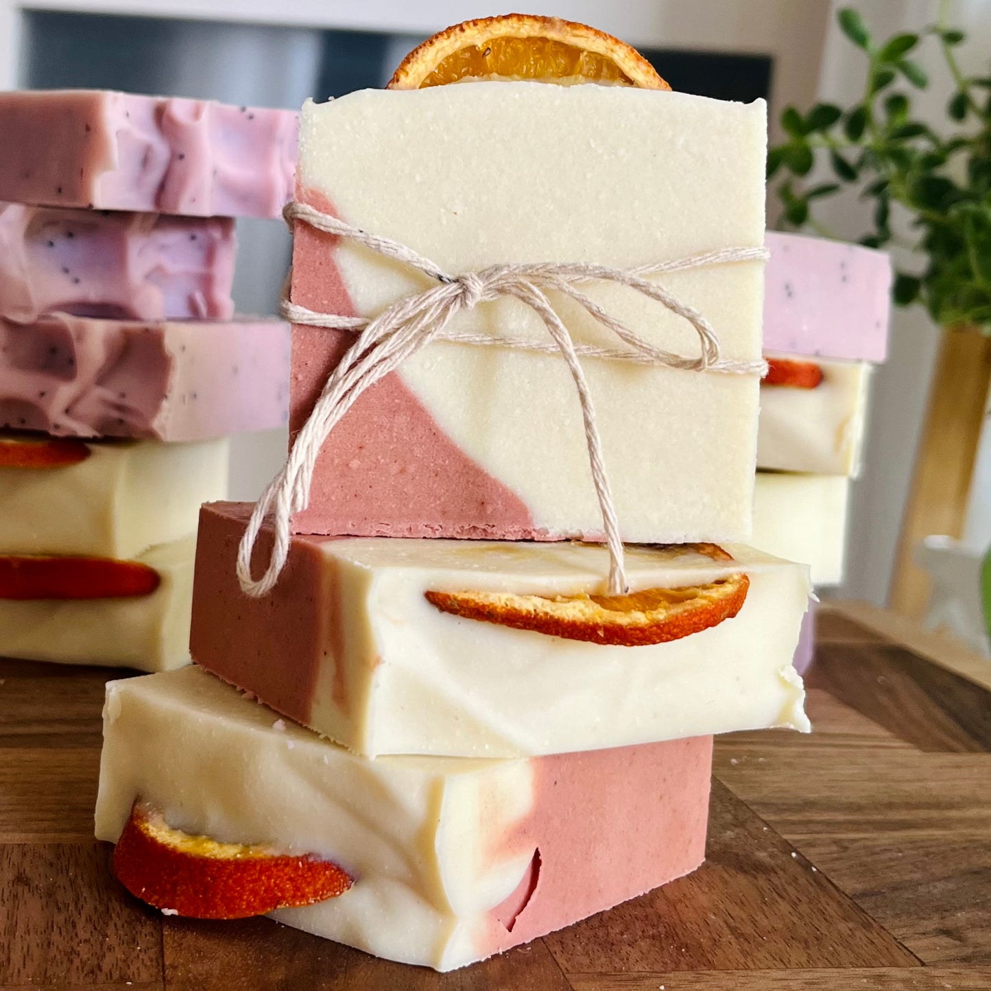 Une pile de trois barres rectangulaires de Savon au lait de coco ZEHRA jojoba et orange faites à la main de La Sultana Savonnerie, avec de magnifiques tourbillons roses et blancs. Chaque barre est surmontée d'une tranche d'orange séchée et nouée avec de la ficelle, promettant un nettoyage luxueux. D'autres pains de savon sont visibles en arrière-plan. Ces barres de savon végane mettent en valeur le savoir-faire naturel des meilleurs savons et produits de soins de la peau du Québec.