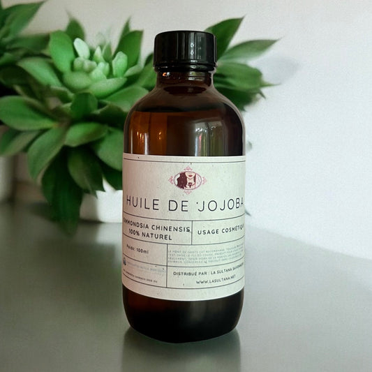 Une bouteille en verre brun étiquetée « Huile de Jojoba » avec un bouchon noir est placée devant une plante succulente verte. L'étiquette indique qu'elle contient 100 ml d'Huile de Jojoba 100% naturelle pour hydrater la peau et les cheveux, distribuée par La Sultana Savonnerie | Savons et soins naturels faits main | Québec.