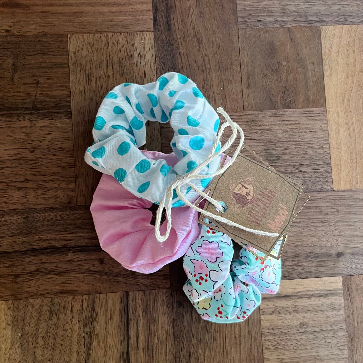 Trois chouchous en tissu reposent sur une surface en bois : un chouchou à cheveux blanc à pois bleus, un chouchou rose et un chouchou blanc à motif floral. Ces beautés faites à la main des "Chouchous à cheveux faits à partir de retailles (paquet de 3)" de La Sultana Savonnerie | Savons et soins naturels faits main | Le Québec se décline en couleurs variables. Une étiquette marron avec le logo de la marque est fixée aux chouchous avec de la ficelle.