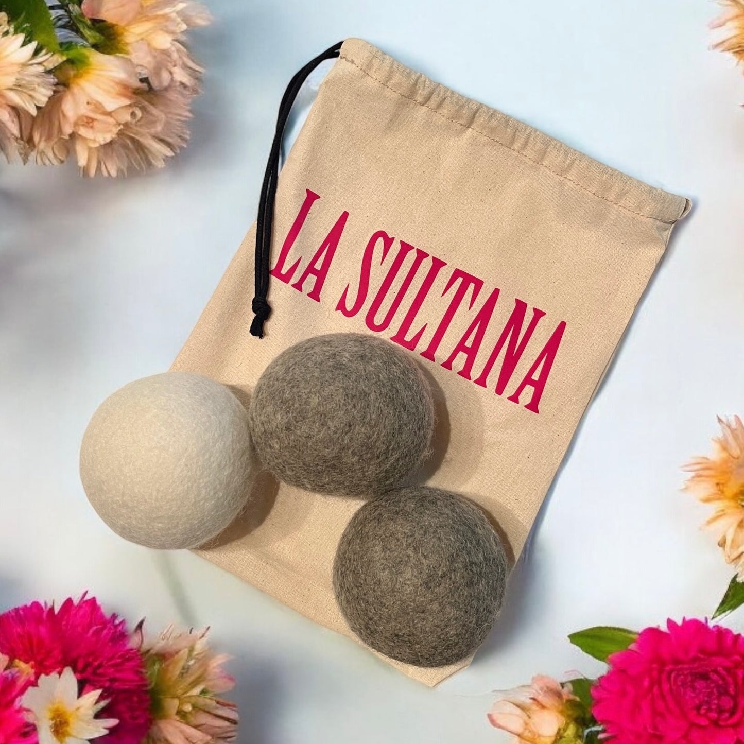 Un sac à cordon beige avec l'inscription "LA SULTANA" en lettres rouges est présenté. Le sac est partiellement ouvert, révélant trois Balles (3) de séchage en laine naturelle en blanc, gris et gris foncé. L'ensemble est entouré de fleurs roses et blanches sur fond bleu clair. Ce produit écologique provient de la Savonnerie La Sultana, connue pour ses savons naturels faits à la main et ses produits de soins de la peau du Québec.