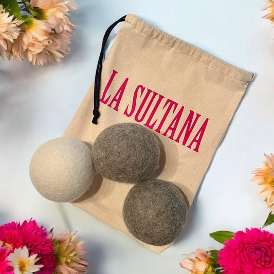 Un sac à cordon beige avec l'inscription "LA SULTANA" en lettres rouges est présenté. Le sac est partiellement ouvert, révélant trois Balles (3) de séchage en laine naturelle en blanc, gris et gris foncé. L'ensemble est entouré de fleurs roses et blanches sur fond bleu clair. Ce produit écologique provient de la Savonnerie La Sultana, connue pour ses savons naturels faits à la main et ses produits de soins de la peau du Québec.