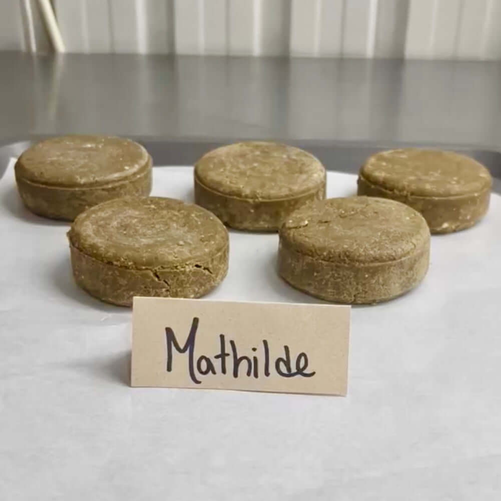 Cinq disques ronds de couleur beige, identifiés comme Shampoing naturel MATHILDE reetha & sidr de La Sultana Savonnerie, sont disposés sur une surface blanche. Devant les barres, il y a un petit panneau en carton avec le nom manuscrit « Mathilde ». Ces options véganes promettent des cheveux doux et brillants. L'arrière-plan semble être un mur carrelé de couleur claire.